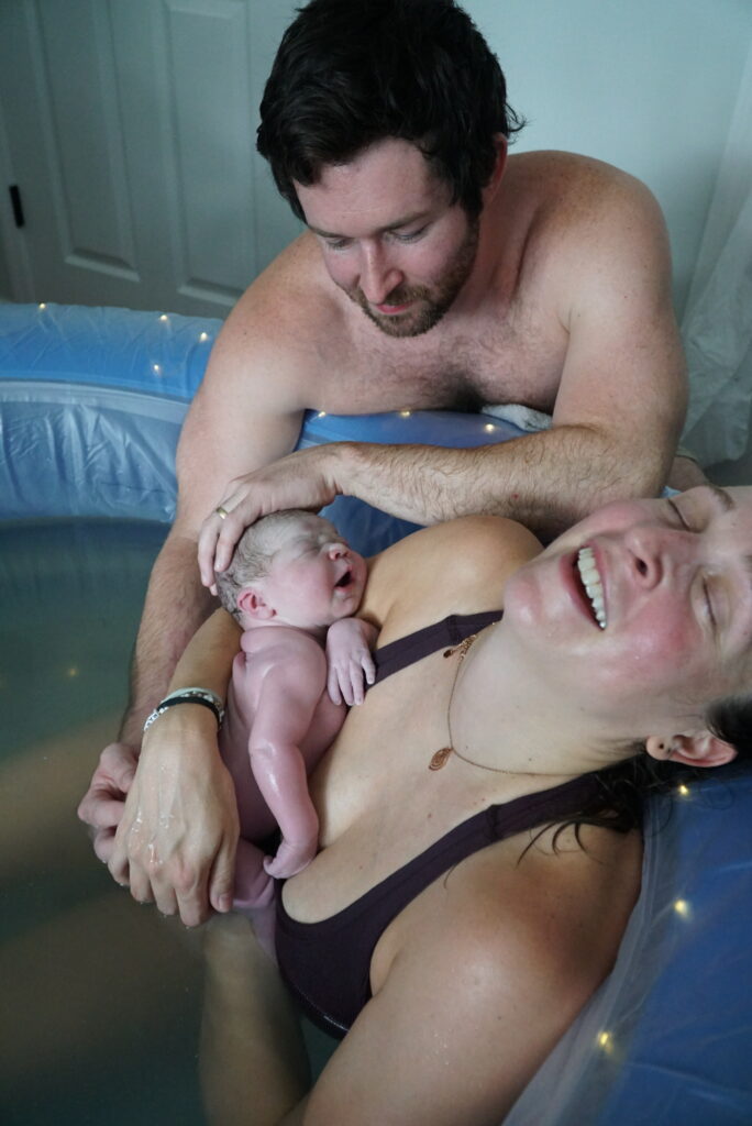 elated-mom-holding-newly-born-baby-in-birth-tub-with-proud-husband-looking-on