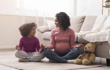 Pregnant-black-mother-and-her-little-daughter-practicing-yoga-at-home